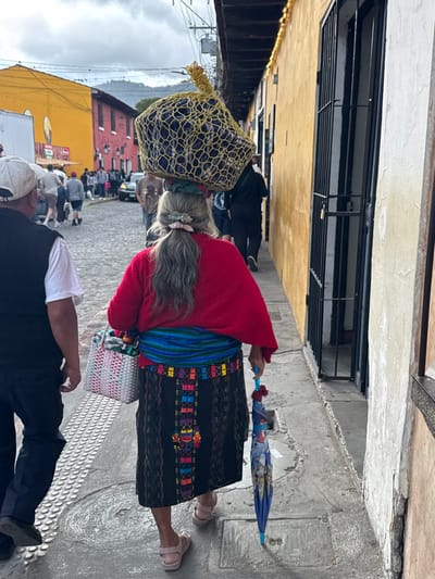 An elderly woman in traditional Guatemalan clothing walks along a cobbled street. She balances a large net bag on her head, filled with dark objects. She wears a red sweater, a dark skirt with colorful embroidery, and a blue-green woven sash. An umbrella is in her left hand and a basket dangles from her right arm. Buildings with yellow and orange facades line the street with blurred people in the background, suggesting a busy market or festival.