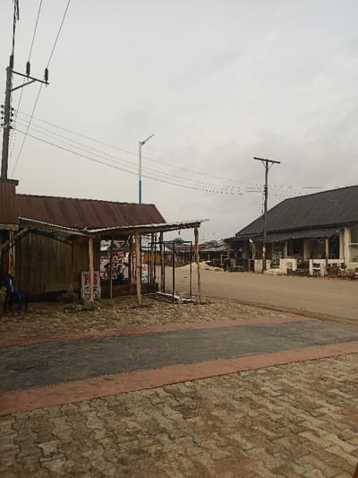 The image shows a street scene in Oghara, Nigeria. The street is paved with red and grey cobblestones, and there is a small wooden building with a corrugated metal roof on the left side of the image. The building is partially enclosed by a wooden structure, and there is a blue plastic chair sitting in front of it. There are several power lines strung between utility poles along the street. The street is empty, and there are no people present. The image was taken during the day, and the weather appears overcast. The background shows several buildings with corrugated metal roofs.