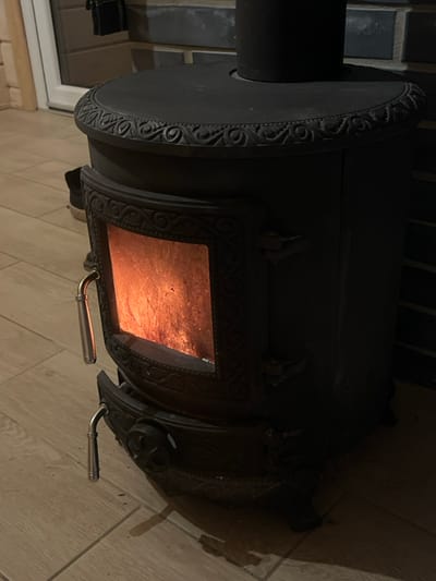 The scene depicts an active, black cast-iron wood-burning stove situated indoors in Petriškė, Lithuania. The stove features a rounded top with ornate scrollwork decoration along its upper edge, and a central black stovepipe extends vertically from its top. A rectangular glass panel on the stove's upper door reveals glowing orange and red embers or flames, indicating active combustion. A polished silver-colored, curved handle protrudes from the left side of this upper door. Below, a second smaller door, presumably for an ash pan or air intake, is also visible, equipped with a similar silver-colored, curved handle.
The stove rests on a light-colored, possibly wooden or tiled floor with visible grout lines. To the immediate left background, a light-colored wooden wall with horizontal planks is visible, alongside a partial white-framed window or door. A dark shoe or boot is partially visible in this area. To the right background, a dark, possibly brick or tiled wall with horizontal grout lines forms the backdrop for the stove. The overall lighting is dim, with the internal fire providing a significant light source and casting shadows on the surrounding floor.