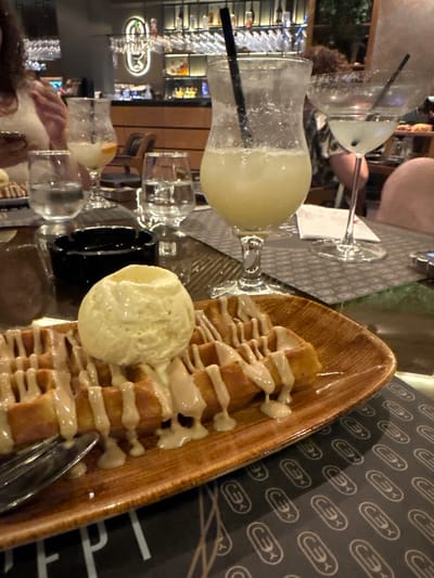 This image captures an indoor dining scene at what appears to be a modern bar or restaurant. The primary focus is a dessert and multiple drinks arranged on a table with a dark placemat featuring a repeating white stylized symbol, alongside the partial white text "CONCEPT." This indicates the location is likely "Concept G Bar & Restaurant" in Strumica, North Macedonia.
In the foreground, a rectangular, reddish-brown ceramic plate holds a golden-brown waffle, seemingly divided into two pieces. The waffle is generously drizzled with a light, creamy, off-white sauce. A single scoop of light yellow ice cream rests on top of the waffle. A silver fork is partially visible on the left side of the plate.
On the table to the left of the dessert is an empty, black ceramic ashtray. To the right of the ashtray, a tall, clear glass contains a light yellow, frothy beverage with a black straw. Further right, another clear glass holds clear liquid (likely water). A margarita-style glass, with a salted or sugared rim, contains a lighter liquid and a black straw, positioned towards the right of the frame.
Several individuals are partially visible: on the far left, a person wearing a light-colored top with dark, curly hair is seen, with an arm holding a phone. On the far right, a bare shoulder and arm are visible, belonging to another patron. In the blurred background, behind the margarita glass, the upper portion of a person's head and hair can be seen.
The background reveals a well-stocked bar area with numerous bottles of liquor on illuminated shelves. Rows of inverted stemware hang from racks above the bar. A prominent, stylized neon-like symbol, resembling the ones on the placemat, is mounted on the wall behind the bar. The overall lighting is warm, contributing to a refined and contemporary atmosphere.
