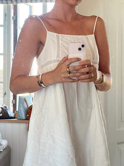 A mirror selfie captures the upper torso and arms of an individual in an indoor setting within Skagen, Denmark. The subject, likely female, wears a white, possibly linen, spaghetti-strap dress featuring a lace-detailed bodice and a simple, thin gold-colored necklace. Both hands grasp a light pink smartphone, equipped with a white textured grip, angled towards the mirror for photography. The left hand displays two gold-colored rings: one wide and one narrow. The right wrist is adorned with a gold-colored watch or bracelet and two additional bangles, one with a white and black pattern and one gold-toned. The background, seen as a reflection, reveals a room illuminated by natural light entering through a window on the left, topped with a light-colored, possibly striped, blind. Below the window, on a surface, several bags are present: a dark one, a light blue one with tan handles, and a reddish-brown one with stud accents. The wall beneath the window is clad in white vertical wood paneling. A white door frame or wall section is visible on the right.