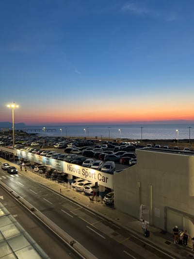 The image captures a parking lot and a coastal road in Cinisi, Italy, during twilight. A vibrant sunset paints the sky with hues of orange, pink, and blue, reflecting on the calm sea. The parking lot is filled with numerous cars, and a large banner displaying "Nuova Sport Car" is visible. A lamppost illuminates the scene, and a road runs alongside the parking lot. Several people are present, some walking with luggage, others with dogs. The overall impression is one of a busy transportation hub at the end of the day, with the beauty of the natural surroundings adding to the atmosphere.