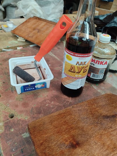 In an outdoor setting, possibly a workshop or yard in the city of Tavatuy, Russia, a close-up shot captures a surface littered with various items related to woodworking or DIY projects. In the foreground, a piece of finished wood with a rich, brown grain rests on a weathered, reddish-brown surface, possibly a workbench. To the left, a white plastic tray holds a small, dark-colored paint roller, partially submerged in a light brown liquid, likely wood stain or varnish. An orange-handled mini paint roller is also visible, its handle extending out of frame.

Dominating the mid-ground are two bottles of wood finishing products. The larger bottle, a clear glass one, contains a dark, viscous liquid, and its label, primarily in Russian, features the word "ЛАК" (LACQUER) in large, stylized lettering, with "ДУБ" (OAK) below it. The label also indicates a volume of 0.5 L and includes product information and warnings. To the right of this bottle is a smaller, clear plastic bottle with a white cap, also containing a similar dark liquid. The labels on both bottles suggest they are intended for wood finishing and protection.

In the background, out of focus, are elements suggesting a cluttered work area, including what appear to be plastic bags, stacks of wood or other materials, and possibly containers of other DIY supplies. The overall scene conveys a sense of ongoing or recently completed manual labor, focusing on the application of a wood finish. The lighting is natural and somewhat diffused, casting soft shadows.