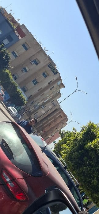 The image depicts a street scene in Berrechid, Morocco, captured from an oblique angle, likely from inside a vehicle. In the immediate foreground, the rear section of a dark red hatchback is visible, including its rear window, wiper, and a portion of the taillight.
On the paved street, in the midground, a male individual is present. He wears a dark blue long-sleeved garment and is positioned between parked vehicles. His head is turned towards the general direction of the camera. Behind him, a dark blue SUV/crossover is parked, followed by a white sedan further down the street, amidst other partially visible parked vehicles.
The background is dominated by a multi-story building, appearing beige or light brown, featuring multiple rectangular windows and external air conditioning units. A prominent sign on this building displays Arabic script identifying it as the "Ministry of National Education." To the left of this structure, a darker-colored building is partially visible.
The environment includes green trees with dense foliage lining sections of the street. Two streetlights with distinctive curved, dual-arm designs extend upwards against a clear, bright blue sky. The scene is illuminated by strong daylight.
