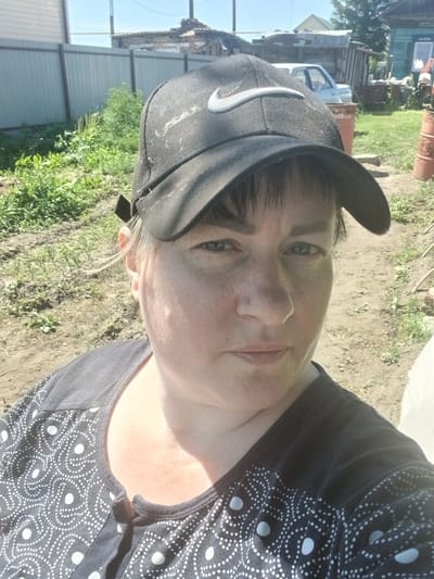 The image is a close-up, outdoor selfie of a woman wearing a black baseball cap with a white Nike logo. She has fair skin, light brown hair, and blue eyes, and she is looking directly at the camera. She is wearing a dark shirt with a white polka dot pattern. The background appears to be a rural or suburban setting, with a wooden fence, some greenery, and outbuildings. There is also a car visible in the background. The overall impression is of a casual, sunny day.
The image is said to be taken in Prokudskoye, Russia. Prokudskoye is a village in the Amur Oblast region of Russia, located in the Far East of the country.