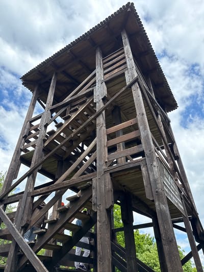 The image shows a low-angle view of a wooden structure, specifically a lookout tower. The structure is brown, and its construction is visible. Some elements are dark due to weathering.
The scene is located in Sremski Karlovci Municipality, Serbia. The image captures the perspective from below, looking up at the lookout tower, partially obscuring the sky. Patches of sky with white clouds are visible through the open framework of the structure. The top of the tower features a decorative roof edge. The tower is built on a steep angle. The stairs up the tower are visible. People are on the stairs.