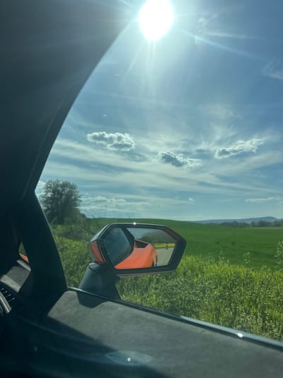 The image depicts a view from inside a vehicle, possibly a car, while driving. The sky is blue with scattered clouds. A side mirror is visible, reflecting an orange vehicle. Green fields line the side of the road, with a distant treeline and hills. The exact location in Blaskovo, Bulgaria cannot be determined from the image.