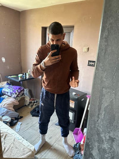 A man is taking a selfie in a mirror. He is wearing a brown hoodie, black pants, and white socks. He is standing in a room with a light brown wall and a hardwood floor. There is a black TV stand with a space heater next to him. There is also a shelf with various items on it. There are also piles of clothes strewn about. The location appears to be indoors but the city of Gromshin, Bulgaria cannot be determined from the information in the image.