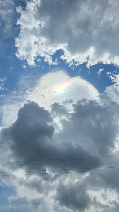 The image captures a vibrant sky scene in Drenkovo, Bulgaria, dominated by a diverse array of clouds under bright daylight. A prominent feature is a large, translucent cloud displaying striking iridescence, with rainbow-like hues of pink, green, yellow, and blue, especially visible in the upper central and left areas. This colorful phenomenon appears around a bright, circular light source, likely the sun, which is partially obscured by the cloud.
The sky is a mix of different cloud types and conditions. To the left and upper left, fluffy white cumulus clouds are interspersed with patches of clear blue sky. In contrast, the right side of the frame features darker, denser gray cloud formations, suggesting a more overcast or impending weather condition in that area. Three thin, dark power lines stretch vertically across the right portion of the image, cutting through these darker clouds. Two small, dark specks, likely birds, are also visible against the cloudy backdrop in the upper right quadrant. The scene depicts a dynamic daytime sky, showcasing natural optical phenomena. No visible text is present.