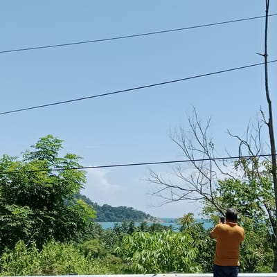 In Layeun, Indonesia, a man in an orange polo shirt and blue jeans stands with his back to the camera, holding a smartphone and appearing to take a photograph. He is positioned in front of a lush, green forest. Beyond the trees, the turquoise ocean is visible, meeting a distant coastline with verdant hills. The sky above is a clear, bright blue, with several dark wires stretching horizontally across the frame. The overall scene captures a moment of observation and appreciation of the natural landscape.