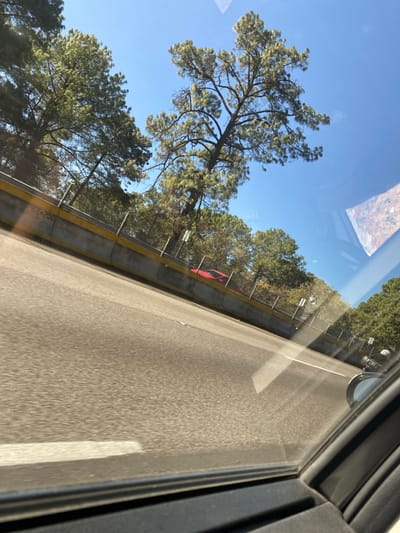 A view from inside a vehicle shows a highway with a concrete barrier and chain-link fence on one side. Trees line the roadside, reaching into a clear blue sky. A glimpse of a red object is visible through the fence. The road surface is asphalt, marked with a white line. The scene is partially framed by the vehicle's window and door frame. Reflections are visible on the window.