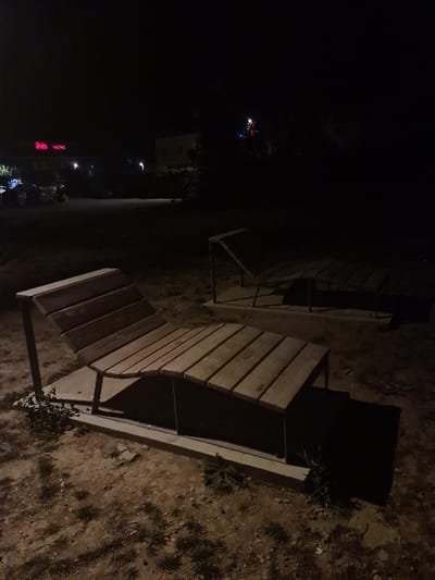 The image displays an outdoor nighttime scene in Les Tourrettes, France. In the foreground, two modern lounge chairs constructed of slatted wood and metal frames are situated on a ground composed of dirt and scattered vegetation. The chair on the left is angled towards the viewer, with its backrest in a reclining position, mounted on a concrete base. A second, similar lounge chair is visible further back and to the right, mostly obscured by shadow. In the background, to the left, a building with an illuminated red neon sign displaying "ibis hotel" is prominent, accompanied by several parked vehicles in a paved lot. To the far right in the background, a cluster of multi-colored lights (red, blue, white) is visible on what appears to be a distant tree or pole. The scene is dimly lit by artificial light sources, casting shadows from the foreground structures to the right. No individuals are present.