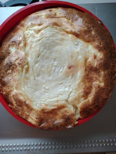 This is a top-down, medium shot of a freshly baked, round cheesecake in a bright red silicone baking dish. The cheesecake has a golden-brown, slightly cracked crust around the edges, with a lighter, creamy filling in the center. The filling appears soft and slightly rippled, with some areas showing a paler, more cooked texture. The baking dish is placed on a gray metal surface that has a textured pattern of raised circles, possibly a baking sheet or the surface of an oven. The background is out of focus, showing a dark, indistinct area at the top and a lighter, lighter gray surface below the textured metal. The lighting suggests it is daytime, and the scene is likely indoors, in a kitchen environment. There is no visible text in the image. The cheesecake appears to be the primary subject, and the overall impression is one of homemade comfort food.