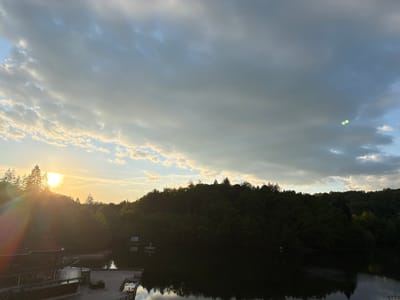 The image shows a lake surrounded by trees at Sovata, Romania. A wooden pier extends into the water in the foreground. The sun is low, obscured by trees on the left, creating a lens flare. The sky is filled with a mix of light and dark clouds. The lake reflects the trees and sky.