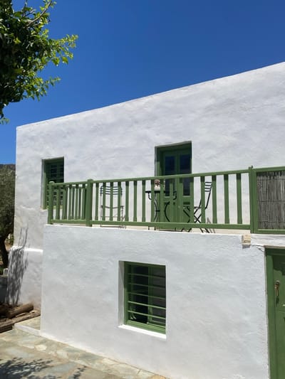 The image displays a two-story building in Πλατύς Γιαλός, Greece, under a clear blue sky. The structure features whitewashed, textured walls. A prominent feature is an upper-level balcony with a green painted wooden railing composed of vertical balusters. On this balcony, a small round green metal table is positioned between two green metal chairs. A light-colored, possibly ceramic, object rests on the table. A green double door provides access to the balcony from the building's interior. To the immediate left of the balcony, a green-framed window with horizontal security bars is visible. Below the balcony, on the lower level, another green-framed window with identical horizontal bars is present. A partially visible green door, from which a key hangs, is located on the far right of the lower level. The ground area in the foreground is paved with flat, irregular flagstones. A tree branch with green leaves extends into the upper left quadrant of the frame, casting shadows. Faint outlines of distant hills are visible on the horizon to the left. The scene is brightly illuminated by direct sunlight, creating sharp shadows. No individuals are present.