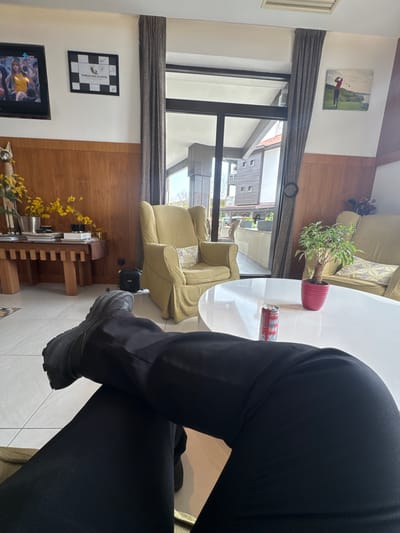The image depicts an indoor scene, likely a living room or lounge, viewed from the perspective of a person sitting at a white, round table. The subject's legs, dressed in black trousers and dark, robust-looking shoes, are prominently featured, crossed at the ankles.

On the white table, a small potted plant in a red pot and a red Coca-Cola Zero Sugar can are visible. The room is brightly lit by natural light streaming through a large window or glass door, which offers a glimpse of residential buildings outside.

The interior decor includes light-colored armchairs and a sofa with patterned cushions, along with prominent wooden wall panels and ceiling beams, and a tiled floor. Various decorative items adorn the space: a framed painting of a golfer in mid-swing on one wall, and below it, a framed piece of art featuring a black and white checkerboard pattern with the text "THRACIAN CLIFFS" and several visible signatures. A television screen, displaying a "9sity" channel logo, is partially visible. A wooden console table, adorned with bright yellow flowers, and a golf bag containing golf clubs are also part of the room's furnishings.

No specific activity beyond sitting is evident for the person. The time of day appears to be daytime due to the natural light. The "THRACIAN CLIFFS" text is a strong location cue, suggesting the setting is in or near the Thracian Cliffs Golf & Beach Resort in Bozhurets, Bulgaria.