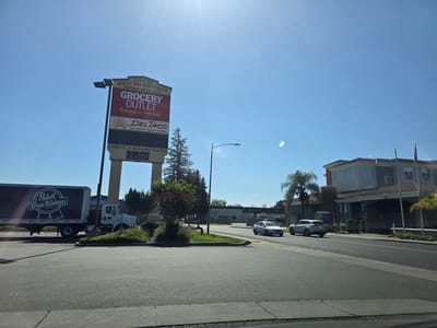 This media file captures a sunny daytime scene in West Sacramento, USA. The image showcases a commercial area with a prominent signpost advertising "CITY OF WEST SACRAMENTO HARBOR BLVD. EXIT" with "GROCERY OUTLET bargain market" and "DELTACO 24 HOUR DRIVE-THRU" listed below. A Pabst Blue Ribbon beer truck is parked on the left side of the frame.
In the background, a Holiday Inn Express hotel is visible, with cars driving on the road in front of it. The sky is clear and blue, indicating fair weather. The presence of palm trees suggests a warm climate. The overall impression is a typical suburban commercial landscape on a bright, clear day. No people are clearly visible in the image, but the scene implies a busy retail and hospitality environment. The "HARBOR-50 BUSINESS CENTER" is also mentioned on the signpost.