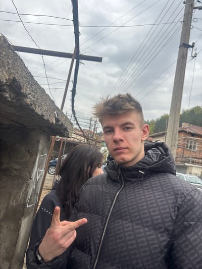 A medium shot, taken from a low angle, shows a light-skinned young man in a dark, patterned jacket, looking directly at the camera. He is standing next to a woman with dark hair, whose face is partially obscured, and who is making a hand gesture. The background includes a weathered concrete wall, a metal structure with wires, an electrical pole with hanging wires, buildings, trees, and a cloudy sky. The location is likely in Bratsigovo, Bulgaria, based on visual elements common to urban areas in that region.