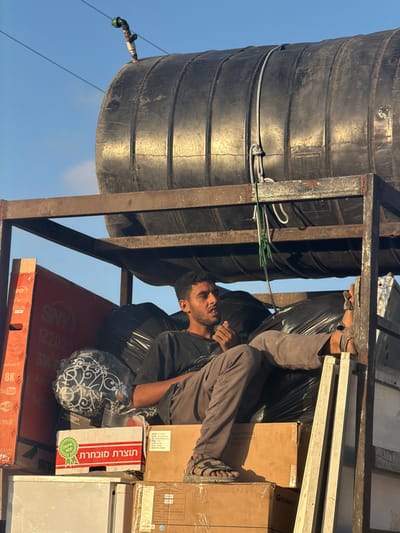 The image depicts an outdoor scene in a-Zawayda, Palestinian Territory. A young adult male, with dark curly hair and a beard, is seated amidst cargo on an elevated platform or structure. He wears a dark short-sleeved shirt, tan trousers, and open-toed sandals. His left leg is extended, resting on a metallic ladder section, while his right leg is bent. His mouth is slightly open, and his left hand is positioned near his chin. He is surrounded by multiple black plastic bags and various cardboard boxes; one box to his immediate left displays Hebrew text "תוצרת מובחרת" (Selected Produce), and a larger red and white box further left is for a "SNY" 8K Android TV. Directly above the male, a large, black, horizontally-oriented, ribbed water tank rests on a rusted dark metal framework. A black and green plastic nozzle protrudes from the top-left section of the water tank, and both white and green ropes are secured to its side, hanging downwards. The background is a clear, light blue sky with two faint utility lines visible in the upper left.
