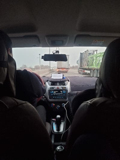 The image depicts the interior of a vehicle, viewed from a rear passenger seat, looking forward through the windshield. The location is Elabad, Uzbekistan.
In the foreground, the backs of two dark-colored car seats are visible, representing the driver's and front passenger's positions. The driver's head and left shoulder are partially visible on the left, and the front passenger's right shoulder is partially visible on the right.
The vehicle's dashboard is gray, featuring a black steering wheel on the left. A rearview mirror is centrally positioned, with a black dashcam mounted below it, obstructing part of the forward view. A smartphone, displaying a map or navigation interface, rests on the dashboard, connected by a white cable that extends towards the center console. The center console includes an infotainment system with a blue display, air conditioning controls, and an automatic gear shifter in the "P" (Park) position. A silver beverage can is visible in a cup holder near the gear shifter.
Through the windshield, a multi-lane roadway under an overcast sky is visible. Several trucks are in view, suggesting traffic. Immediately ahead, a white box trailer truck is present. To the right, another truck is seen carrying a large, green, lattice-like structural component. Distant trees and roadside infrastructure line the horizon. The overall impression is that of a journey in traffic on a highway during daytime with muted lighting.