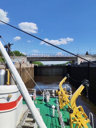 The image captures a scene in the city of Uglich, Russia. It features a view from the deck of a boat, possibly a tugboat, located within a river lock. The boat has a green deck and a white structure with a red stripe. There are yellow cranes visible on the deck. The lock walls are made of concrete, with water filling the space between them. A bridge spans the lock, and trees are visible on either side of the riverbank. The sky is blue with scattered white clouds.