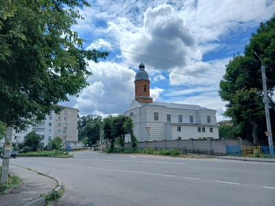 The image shows a street scene in Stryzhavka, Ukraine. A multi-lane asphalt road curves through the foreground. To the left of the road, a sidewalk is visible, with a manhole cover embedded in it. Beyond the sidewalk, there is a large, mature tree with green foliage, partially obscuring the sky. In the background, on the right side of the road, a distinctive building with a red brick tower topped by a grey dome stands prominently. This building has a white facade with multiple windows and decorative elements. It is enclosed by a grey brick wall topped with a black metal fence. To the left of this building, a tall, light-colored apartment building with many windows is visible. The sky is partly cloudy, with large white cumulus clouds against a blue background. There are power lines crossing the scene, supported by poles. Several trees are scattered throughout the background, adding to the greenery of the environment. A blue and yellow fence is situated along the perimeter of the property with the tower building. A traffic sign indicating a curve to the right is visible near the road.