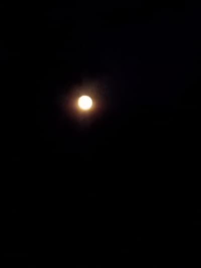 The image depicts a dark night sky with a bright, luminous full moon. The moon is positioned slightly off-center to the left and is surrounded by a soft halo of light, indicating it may be partially obscured by thin clouds or atmospheric haze. The background is entirely black, suggesting the absence of other visible celestial bodies or terrestrial light sources. The photograph was taken in Yampil, Ukraine, likely during nighttime hours. There are no discernible people, objects, or actions depicted in the scene, focusing solely on the celestial phenomenon of the moon.