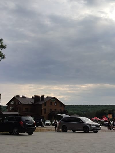 The image captures an outdoor scene with a hotel building in the background, situated in the village of Shcherbani, Ukraine. The hotel is constructed of dark wood and features multiple windows and chimneys on its roof. A green sign displaying "HOTEL" with four stars is visible on the hotel's facade. In the foreground, several vehicles are parked on a paved surface. These include a black SUV, a grey minivan with its trunk open, and other cars in the middle ground. Several people are visible near the minivan and in the background near tents. The sky is overcast with a mix of grey and lighter clouds. The environment includes trees and hills in the distance.