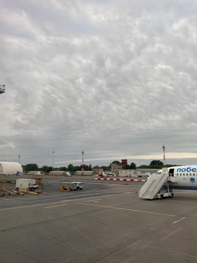 The image shows an airport apron under a cloudy sky. An airplane with "Победа" (Pobeda) written on its side is parked, with a set of stairs attached. Various ground vehicles and equipment are visible on the apron, including a small truck and several other utility vehicles. In the background, there are buildings and trees. The location is identified as Башкултаево, Russia.