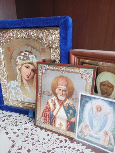 The image depicts a collection of religious icons and a postcard displayed on a crocheted doily. The largest icon, on the left, is framed in gold and features a depiction of the Virgin Mary. To its right, a smaller icon shows Saint Nicholas, holding a book and extending his hand in blessing. In the foreground, a postcard displays an image of Jesus Christ with angels. The objects are placed on a doily, which is on a surface that appears to be a table. The background consists of a wooden shelf. The location is specified as Maryanskaya, Russia.