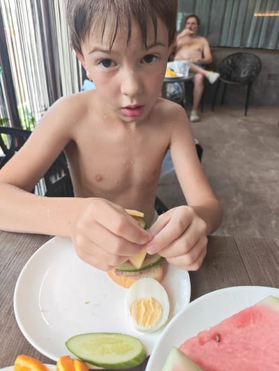 A young boy with wet hair and skin is making a sandwich at a table. He is holding a piece of cheese and placing it on a piece of bread with sliced cucumber and what appears to be a spread. On the plate in front of him are a hard-boiled egg, sliced cucumber, and a slice of watermelon. In the background, an adult male is sitting at another table, eating. The scene appears to be outdoors, possibly at a cafe or restaurant, with a patterned curtain in the background. The overall mood seems relaxed and casual. The time of day is likely daytime, given the lighting. There is no visible text in the image.
