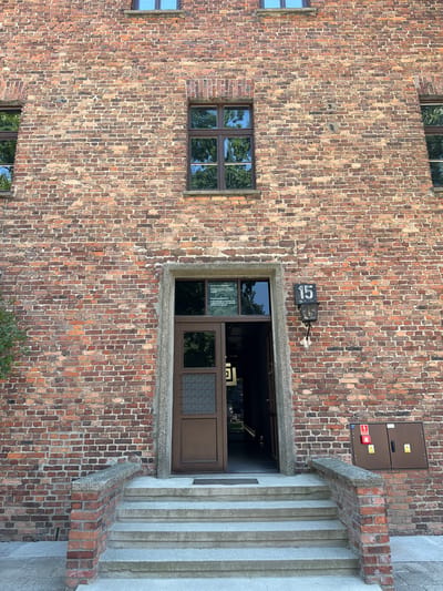 This media file is a photograph of the exterior of a brick building, likely part of a historical site given the location context of Oświęcim, Poland. The building's facade is constructed from a variety of red and brown bricks, laid in a stretcher bond pattern. Several windows are visible, some appearing to be modern with dark frames and glass reflecting trees. One prominent window is larger and has a dark frame with multiple panes, some of which are frosted or textured. Above this window, a dark sign with the number "15" in white digits is mounted on the brick wall, along with what appears to be a small black box with a handle. Further to the right, a concrete staircase with vertical grooves leads upwards alongside the brick wall. Above the large window, a metal cabinet with two doors is mounted on the wall. This cabinet has warning symbols and what looks like a fire alarm. The scene suggests an outdoor setting with daylight, though the time of day and specific weather conditions are not definitively discernible. There is no visible indication of people or any specific activity taking place. The overall impression is one of a historical and possibly somber location, indicated by the brick architecture and the presence of the numbered sign.