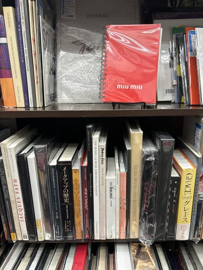 The image shows a bookshelf filled with books. The left side of the shelf has a red notebook with the word "MIU MIU" on it. The right side of the shelf has a stack of books, all different sizes and colors. The topmost book on the right side of the shelf is titled "GRACE." The books below it are titled "Dessous," "111 DARLINGS," "Making Faces," "The Model As Muse: Embodying Fashion," "Modern Souls," "what is beauty?," "ベーシックな装い," "Erwin Blumenfeld," "Jacques Fath," and "UN ART AUTRE." The location of the bookshelf is in Chiyoda City, Japan, which is located at 35.696 latitude and 139.76 longitude.