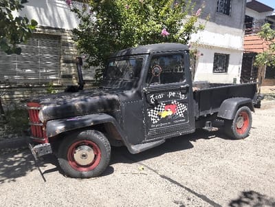 A vintage, black pickup truck with red-rimmed wheels is parked on a street. The truck has a weathered appearance with visible wear and tear. The driver's side door features a graphic of a checkered flag and a cartoon character. The truck's hood is also black and rusty, with a vertical exhaust pipe. The background includes houses and a tree with pink flowers.