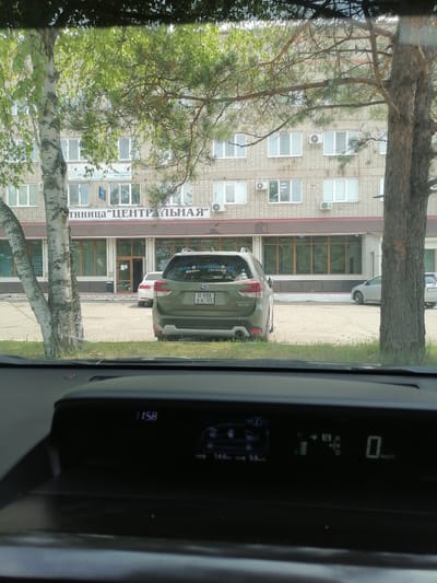 The image shows a view from inside a car looking out at a building and a parking lot. The building is a multi-story brick structure with numerous windows and several air conditioning units. A sign on the building reads "ТИНИЦА ЦЕНТРАЛЬНАЯ" which translates to "CENTRAL CLINIC". In the foreground, a green Subaru Forester is parked. The car has a license plate that reads "O 938 XK 125". There are other cars parked in the lot, including a white car and a silver car. Trees are visible in front of the building and to the sides of the frame. The interior of the car shows a digital dashboard display with the time "11:58" and other indicators. The scene appears to be in the city of Lesozavodsk, Russia, as indicated by the presence of Russian text on the building and the license plate format.