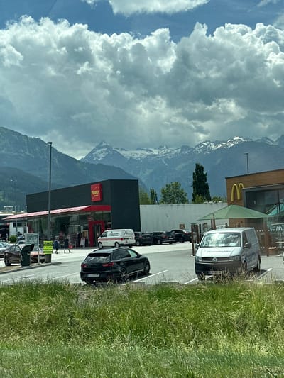The image shows a street view in Zell am See, Austria, likely during the daytime. In the foreground, a grassy verge lines a paved area with several parked cars, including a black Audi and a white Volkswagen van with "Hasenbichler" written on its side. Further back, a Penny Market store is visible with its distinctive red and black signage. Adjacent to it is a McDonald's with its iconic golden arches.

Behind the buildings, a majestic mountain range dominates the background, with some peaks still showing snow despite the greenery and cloudy sky. The sky itself is a mix of blue and heavy, dramatic clouds, suggesting possible impending rain or a recent shower. The overall scene depicts a commercial area set against a stunning natural landscape.

Visible text includes "PENNY MARKET", "McDonald's", "Hasenbichler", and a sign that appears to read "WELTCUP 15.-21. JUN)", possibly advertising an event. The presence of the Penny Market and McDonald's suggests a shopping or dining area. The cars and people suggest normal daily activity. The weather appears dynamic, with bright sunlight intermittently breaking through the clouds, casting varying light on the scene.