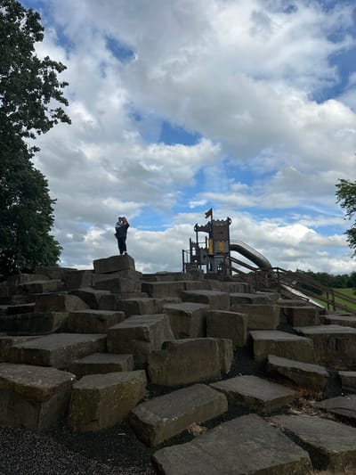 A low-angle, full shot captures a naturalistic playground area against a dynamic cloudy sky. In the foreground, large, rough-hewn grey stones are arranged in a staggered, tiered formation, creating a challenging, rocky landscape for play. A person, likely an adult, stands on one of the higher stones, holding a young child. The adult is wearing dark clothing and the child is wearing a light-colored top. They appear to be posing for a photo, with the adult waving. Behind them, to the right, is a modern playground structure designed to resemble a castle, complete with turrets and a large, silver slide. Wooden safety barriers and a ramp lead up to the slide. The background shows a lush green landscape with trees and a clear blue sky peeking through dramatic white clouds. The scene is set in a park or outdoor recreational area. The location is given as a playground in the city of Wakefield, United Kingdom.