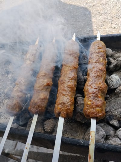 Four skewers of meat kebabs are grilling over coals, emitting a visible plume of smoke. The kebabs are elongated and cylindrical, with a textured, browned surface indicating they are cooked. They are positioned over a black metal grill filled with charcoal. The background shows a rough, gravelly surface, possibly a path or ground outside. The scene appears to be set outdoors, under natural light. The image does not contain any people or explicit references to the city of Ala-Buka, Kyrgyzstan.