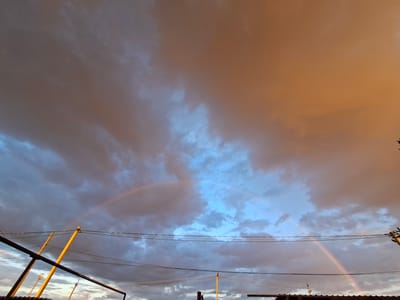 The image depicts a sky filled with dramatic clouds, exhibiting a transition of colors from a warm orange hue on the right to a cooler blue on the left. Two faint rainbows are visible arching across the sky. In the foreground, electrical wires are strung between poles, with some metallic structures and what appears to be rooftops visible at the bottom. The scene suggests a post-rain or approaching storm scenario, with the setting or rising sun casting light on the clouds. The location is identified as Novostroyka, Russia.