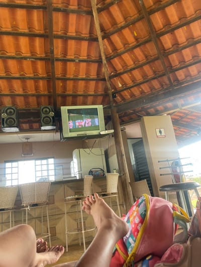 The image depicts an interior view of an open-air establishment, identified as being in Pirenópolis, Brazil. The upper frame is dominated by a vaulted ceiling constructed of reddish-orange clay roof tiles, supported by exposed dark wooden beams and rafters. Scattered yellow particulate matter is visible on the roof tiles.
Centrally, a silver-grey cathode-ray tube (CRT) television is mounted on a wooden beam, displaying an image with a female face on the left and a male face on the right, consistent with video playback. To the left of the television, two black, two-way loudspeakers are mounted on a shelf. Below these, a light brown bar counter with a geometric pattern along its front extends across the mid-ground. Behind the bar, a white, two-door refrigerator is visible, along with a wooden, drum-shaped pendant light fixture hanging from the ceiling. Further left, a multi-pane window allows bright natural light into the area. Several light-colored, wicker-style bar stools with patterned bases are positioned in front of and around the bar counter.
On the right side of the frame, a light brown wall features a rectangular sign with a black female pictogram and the Portuguese word "FEMININO," indicating a women's restroom. In the foreground, two pairs of bare human feet and lower legs are visible, positioned as if the individuals are in a relaxed, reclined posture. A pink backpack with a multi-colored, abstract pattern and a yellow zipper is partially visible in the lower right corner, resting near one of the feet. The scene suggests a casual daytime environment where individuals are relaxing and watching television.