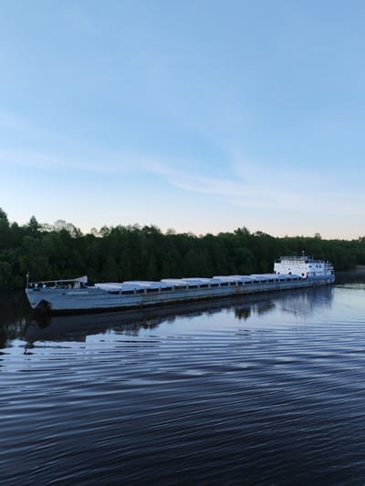 This media file captures a long, grey cargo barge moving or moored on a calm river. The barge has numerous large, light-colored covers over its cargo holds and a multi-deck white cabin structure at its stern. Dense green forest lines the distant riverbank, creating a natural backdrop.
The sky is a clear, light blue with subtle wisps of clouds, suggesting either early morning or late evening due to the soft, even light. The river's surface is smooth with gentle ripples, reflecting the sky and the trees. The overall atmosphere is peaceful and serene.
Visible white text on the bow of the barge, as well as on the side of the stern cabin, reads "НОВОКУЙБЫШЕВСК" (Novokuybyshevsk), likely indicating its name or homeport. No people are visible in the image. The setting appears to be a natural waterway, consistent with the location of Volokov Most, Russia.