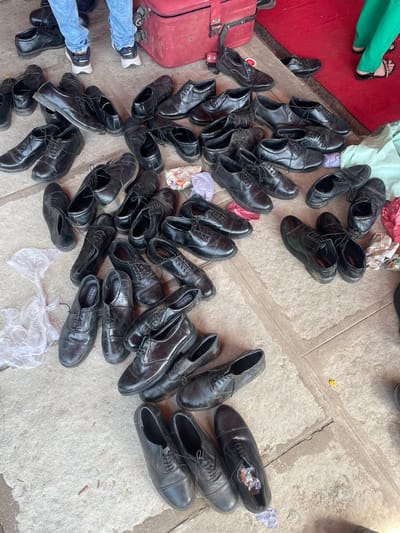 A large pile of black leather dress shoes is scattered on a concrete surface. Some shoes contain crumpled paper or tissue inside. A red suitcase is visible to the left, partially covered with shoes. Two people's legs are seen; one wearing green pants and sandals, the other wearing jeans and sneakers. The shoes appear worn, suggesting they are used or for sale. The scene, likely a marketplace or street vending area, possibly takes place in Anajpur, India.
