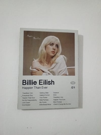In the city of Al Haouafate, Morocco, a rectangular print, designed in the style of a music album cover poster, is affixed to a plain white wall. The print is vertically oriented and features a split design. The upper section contains a photographic portrait of musician Billie Eilish, depicted from the chest up, with platinum blonde hair and bangs. She looks slightly to her right (viewer's left) with a neutral expression, wearing a light-colored, possibly white, garment that is partially pulled down, exposing her shoulders. The background behind her is a warm, solid beige color. The text "Happier Than Ever" in a script font is present in the upper left corner of this photographic area.

The lower section of the print has a solid white background with black text. The artist's name, "Billie Eilish," is prominently displayed in a large, bold font, followed by the album title, "Happier Than Ever," in a slightly smaller, bold font. Below this, a multi-column tracklist is visible, containing song titles such as "Therefore I Am," "Everybody Dies," "Getting Older," "Halley's Comet," "Your Power," "Oxytocin," "NDA," "Male Fantasy," "My Future," "Lost Cause," "OverHeated," "Billie Bossa Nova," and "I Didn't Change My Number." In the bottom right corner of this section, a stylized globe-like icon is accompanied by the numerical designation "/21." The print itself appears slightly raised from the wall, casting a faint shadow along its right edge. The white wall surface is unadorned.
