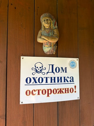 The image displays a rustic wooden wall with a decorative carving of a Native American figure at the top. Below the carving, a white rectangular sign is affixed. The sign features a skull and crossbones symbol in blue, followed by the Russian text "Дом охотника" (Hunter's House) in large blue letters. Below this, in larger, bolded red and yellow letters with a yellow outline, is the word "осторожно!" (caution!). To the right of the main text on the sign, there is a small circular sticker with a blue border and a light blue center containing text and small images, possibly indicating a certification or approval. The environment appears to be outdoors, suggested by the wooden structure and natural lighting. The location is identified as Yagul, Russia, though the specific context of the sign's placement within the city is not provided by the image alone.