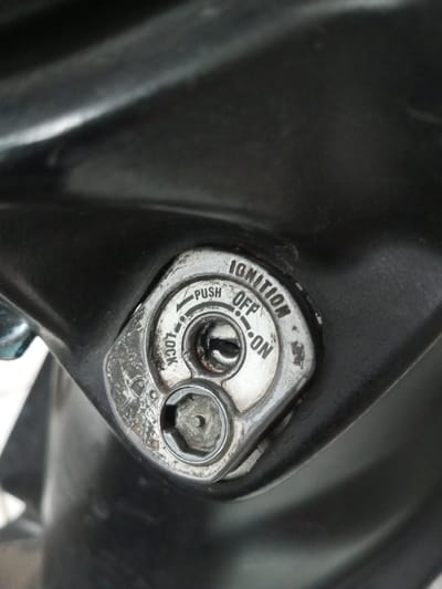 Close-up image shows a motorcycle ignition switch. The switch is silver with black lettering indicating "IGNITION," "OFF," "ON," and "LOCK." Arrows are used to show the direction of the switch to engage these functions. The keyhole is located in the center of the upper portion of the switch. Below the keyhole is a circular hex key. The switch is mounted on a black surface. The condition of the switch indicates it has been used.
