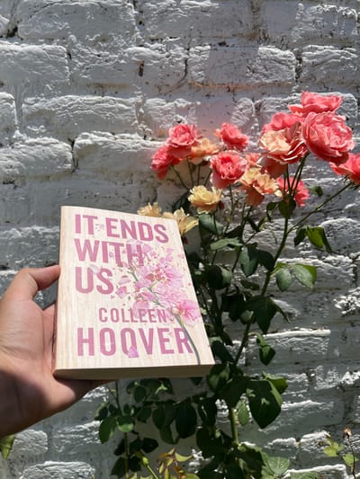 The image shows a human left hand holding a paperback copy of the book "IT ENDS WITH US" by Colleen Hoover. The book's cover features a light, wood-grain texture with the title and author's name in pink typography, adorned with an illustration of scattered pink, orchid-like flowers. The hand grasps the bottom-left edge of the book. In the background, a vibrant rose bush displays numerous blooms in shades of coral pink, peach, and pale yellow, interspersed with green leaves and stems. Behind the roses and the book is a textured white-painted brick wall, showing the uneven surfaces of the bricks and mortar. The scene is outdoors and brightly lit by direct sunlight, evident from the strong highlights and sharp shadows cast by the hand on the book, and by the foliage and blooms on the wall. The exact location in the city of Kabal, Pakistan, cannot be determined from the visual information present in the image.