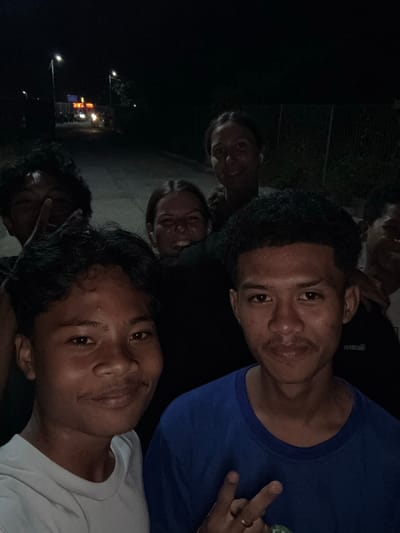 The image depicts a group of young people outdoors at night, possibly in Labuhan Lombok, Indonesia, given the context. Several individuals are visible, with varying degrees of focus. The lighting suggests a street or open area, with a few light sources casting illumination. In the background, a street with a few vehicles and lights can be seen, along with what appears to be a fence. Some of the individuals are smiling and looking towards the camera, while others are partially obscured. The overall impression is a candid snapshot of friends gathered at night.