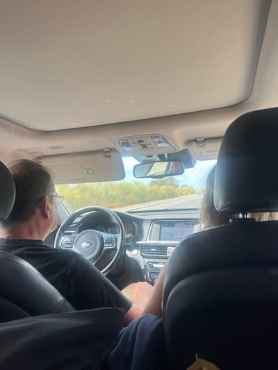 The image depicts the interior of a Kia vehicle, observed from the rear passenger seat, traveling on a road in or near Ayamonte, Spain. A male driver, wearing a dark short-sleeved shirt and glasses, is positioned in the left-hand driver's seat, gripping the steering wheel. His right leg is visible, wearing light-colored shorts. To his right, in the front passenger seat, is a person whose back is visible, characterized by long, light brown hair. This passenger's left leg, clad in dark blue fabric, is positioned close to the driver's right leg.
The car's interior features a beige headliner, a sunroof, and an overhead console with light controls. A lowered sun visor is visible on the driver's side. The dashboard includes a multimedia screen displaying a map or navigation interface, and the rearview mirror reflects a partial image of a person or object behind the front seats.
Through the windshield, a multi-lane asphalt road is visible, flanked by dense green and light brown vegetation under a clear, bright sky. Another vehicle is distantly visible ahead on the road.