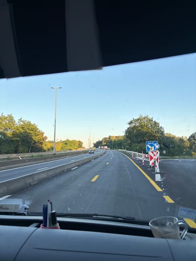 This media file depicts a view from inside a vehicle, looking out onto a highway in Meise, Belgium. The highway appears to be under some form of construction or maintenance, with temporary barriers and directional signs visible. The road itself is asphalt with yellow lane markings. The sky is clear and blue, indicating daytime, likely late afternoon or early evening due to the warm light illuminating the trees.

In the foreground, a portion of the vehicle's interior is visible, including a dark dashboard and a holder with pens or markers. The scene is dominated by the highway stretching into the distance, flanked by lush green trees on either side. Several streetlights are also present along the roadside. In the mid-ground, traffic signs are erected, including a blue directional sign with arrows and a hazard warning sign with red and white stripes. Some vehicles are seen on the highway, suggesting active traffic flow. The overall impression is one of travel on a sunny day with ongoing roadwork. No people are clearly identifiable. No visible text is legible.