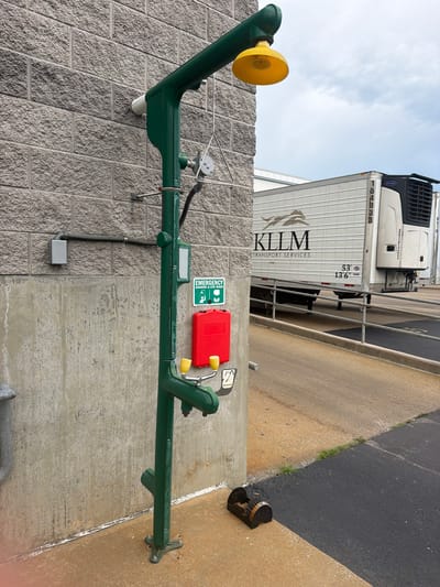 In Murfreesboro, United States of America, an outdoor emergency shower and eye wash station is positioned against a textured gray brick wall and a section of rough concrete. The station, painted forest green, features a shower head with a yellow canopy, an eye wash faucet with two yellow handles, and a bright red emergency box mounted on the structure. Above the red box, a green sign indicates "EMERGENCY SHOWER & EYE WASH" with pictograms. To the left of the station, a metal conduit runs horizontally along the wall, connecting to a gray electrical box. In the background, a white semi-trailer truck with "KLLM TRANSPORT SERVICES" branding is parked on an inclined concrete ramp. The ground in the foreground is a mix of tan concrete and black asphalt, with a small patch of grass. A black boot brush with metal bristles rests on the ground near the base of the shower station. The sky above is overcast with clouds.